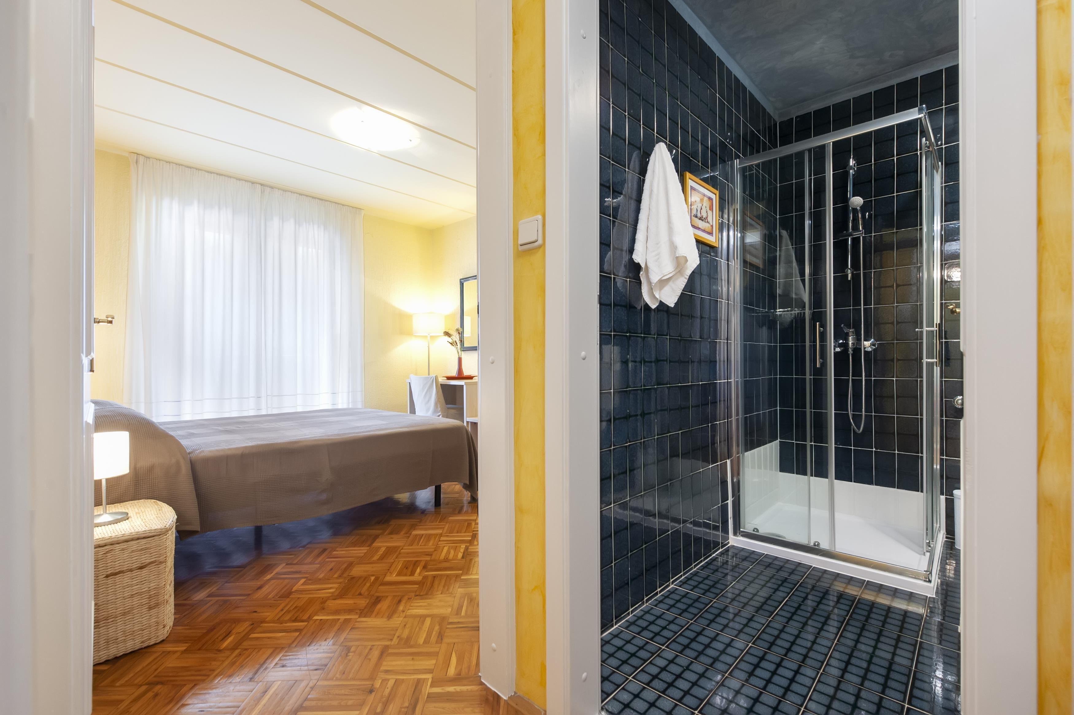 A hotel room with a bed, bedside table, lamp, and a shower area with black tiles.