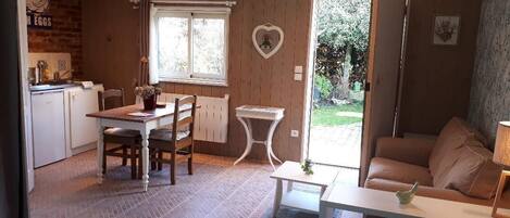 A cozy room with a bed, a dining table with chairs, a small white table, a sofa, and a window with a heart-shaped decoration.