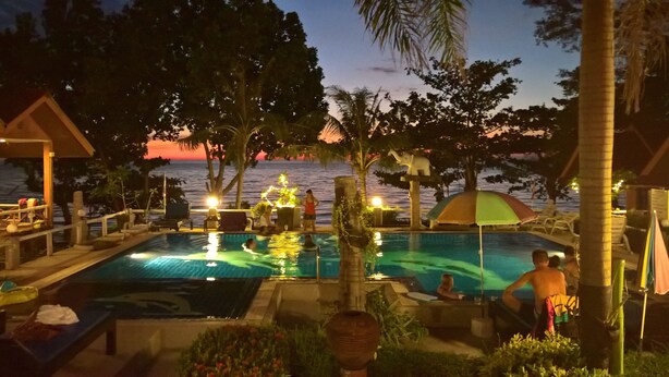 A pool area with lounge chairs and umbrellas, surrounded by palm trees and a view of the sea at dusk. - Penny Bangalow Resort (Ko Chang)