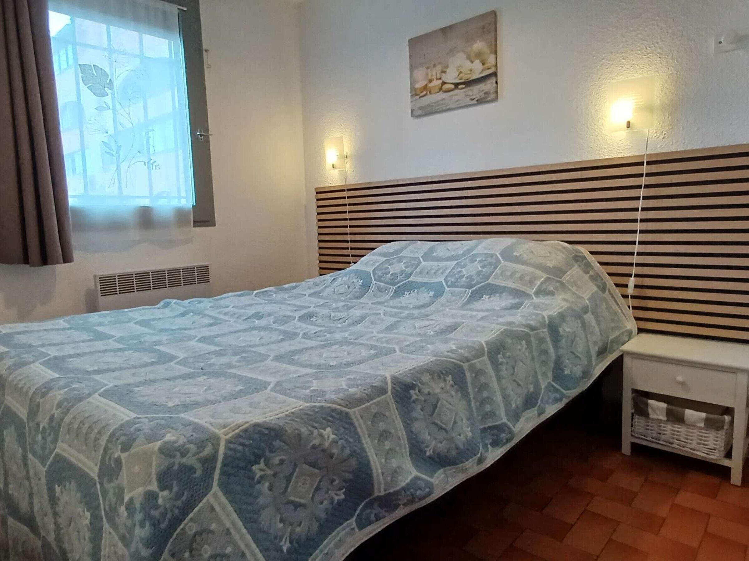 Une chambre à coucher comprenant un lit, une table de chevet, une lampe fixée au mur et une fenêtre avec des stores.