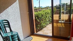 A balcony with a chair, a glass door, and a view of greenery and a palm tree.