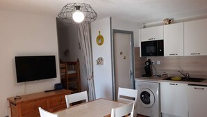 A compact kitchen with a microwave, sink, and washing machine. There is a wooden dining table with chairs, a TV mounted on the wall, and a door leading to another room.