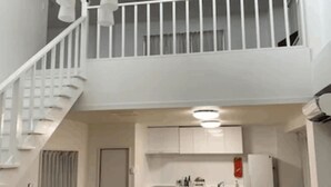 En modern lägenhet med trägolv, en svart lädersoffa, en matplats med kök och en vit trappa.