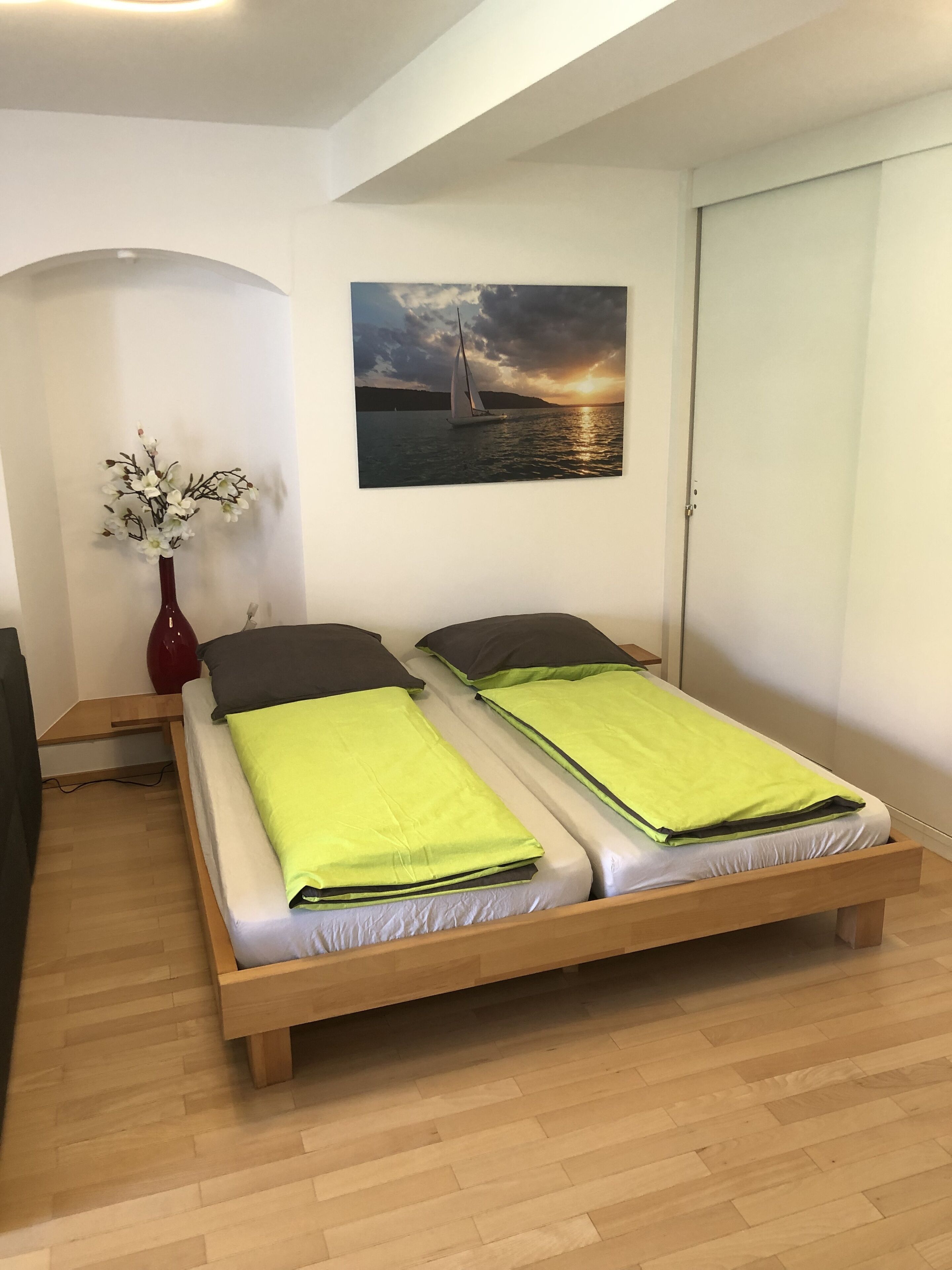 A bedroom with two beds, a wooden floor, a painting of a sailboat on the wall, and a vase with flowers.