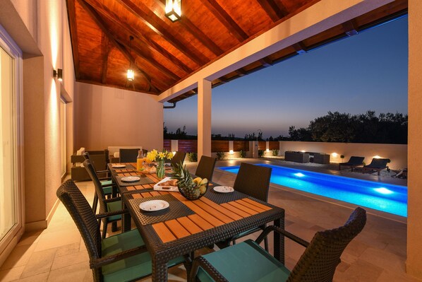 An outdoor dining area with a pool view, featuring a wooden ceiling and outdoor furniture.