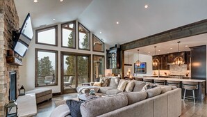 Un salón amplio con un sofá modular grande, una chimenea de piedra y una zona de cocina con electrodomésticos modernos e iluminación colgante.