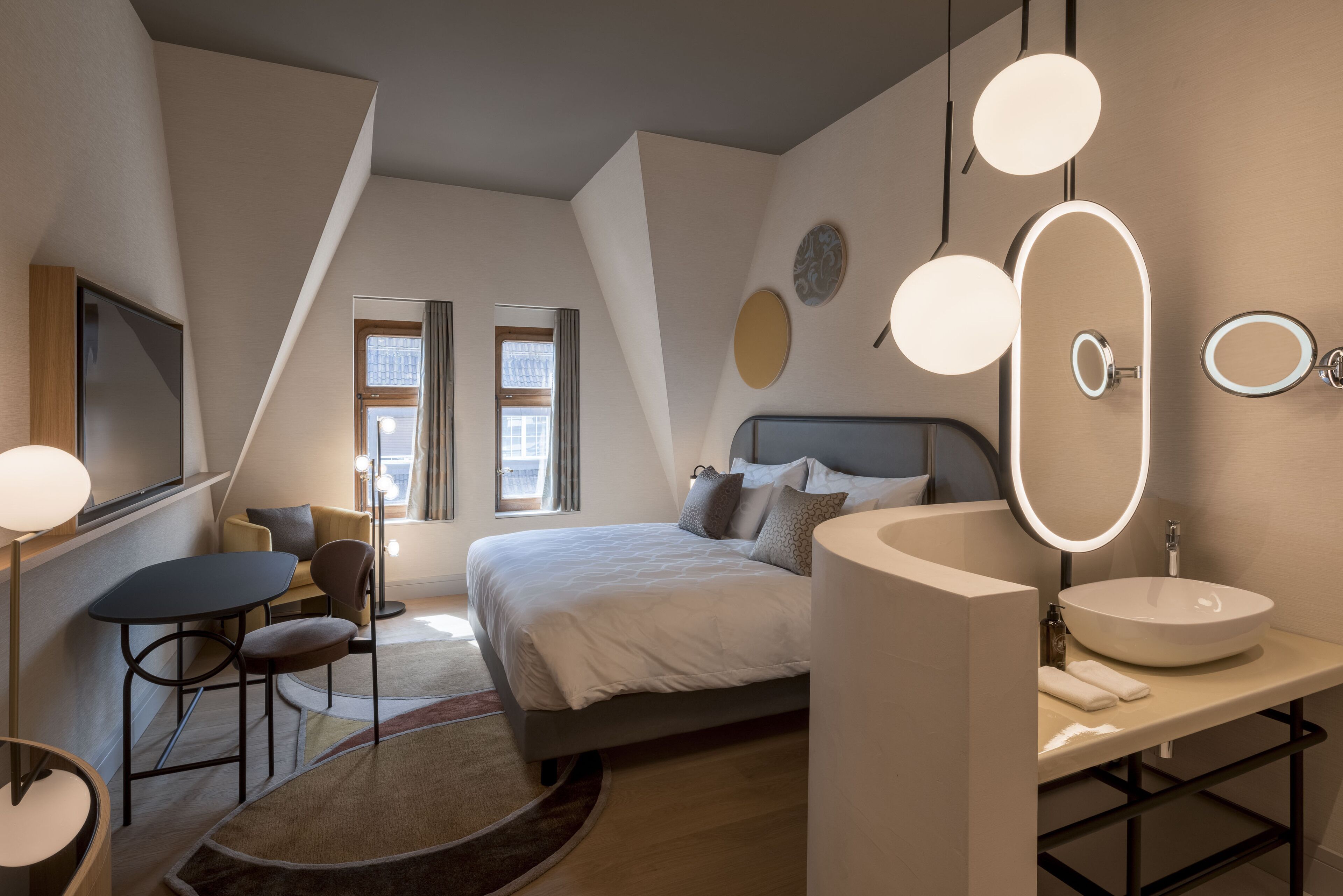 Una habitación de hotel moderna con una cama, mesita de noche y un espejo redondo grande. Hay una pequeña zona de estar con una silla y una mesa.