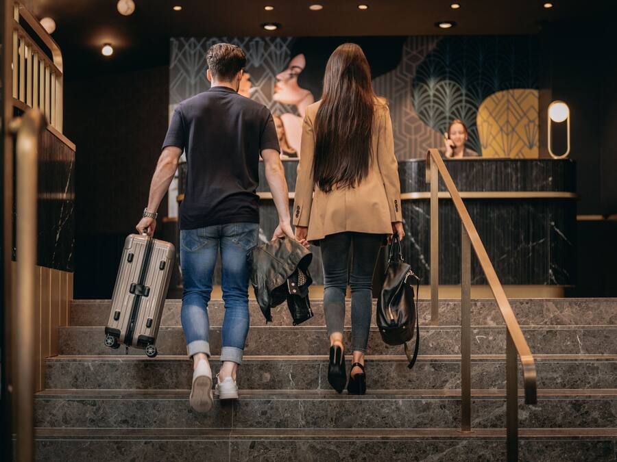 A man and a woman walking down a staircase with a handrail, holding a suitcase and a handbag.