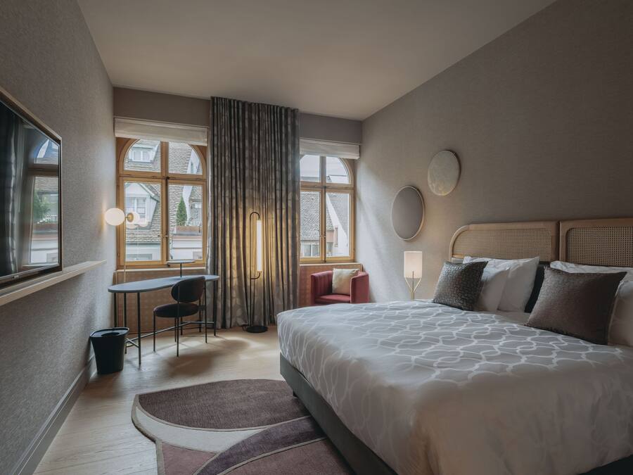 A modern hotel room with a large bed, a desk, and a chair. There is a window with curtains, a round mirror on the wall, and a small trash bin.