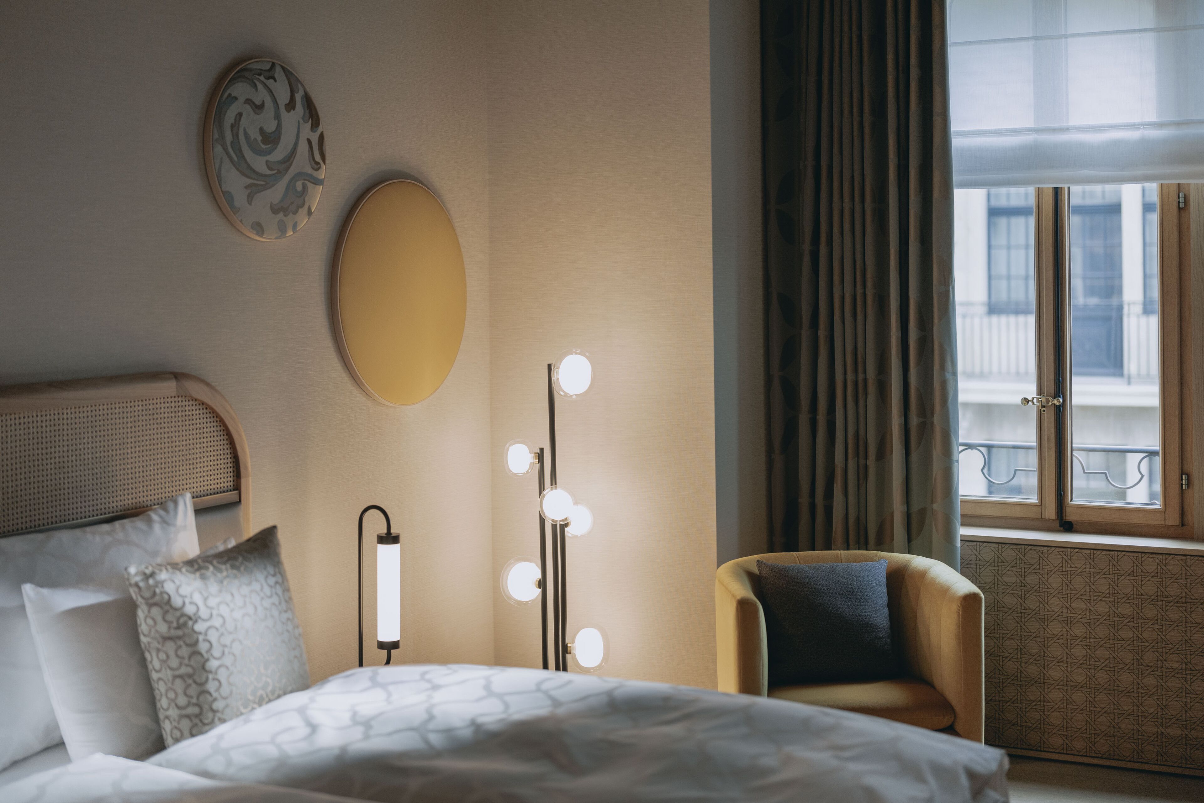 Ein Hotelzimmer mit Bett, Sessel, Schminktisch mit Beleuchtung, großem Spiegel und einem Fenster mit Vorhängen.