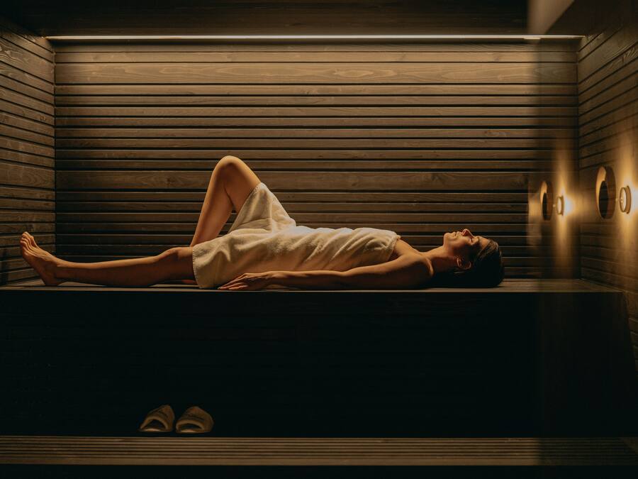 A person lying on a bench in a wooden room with built-in benches and wall-mounted lights.