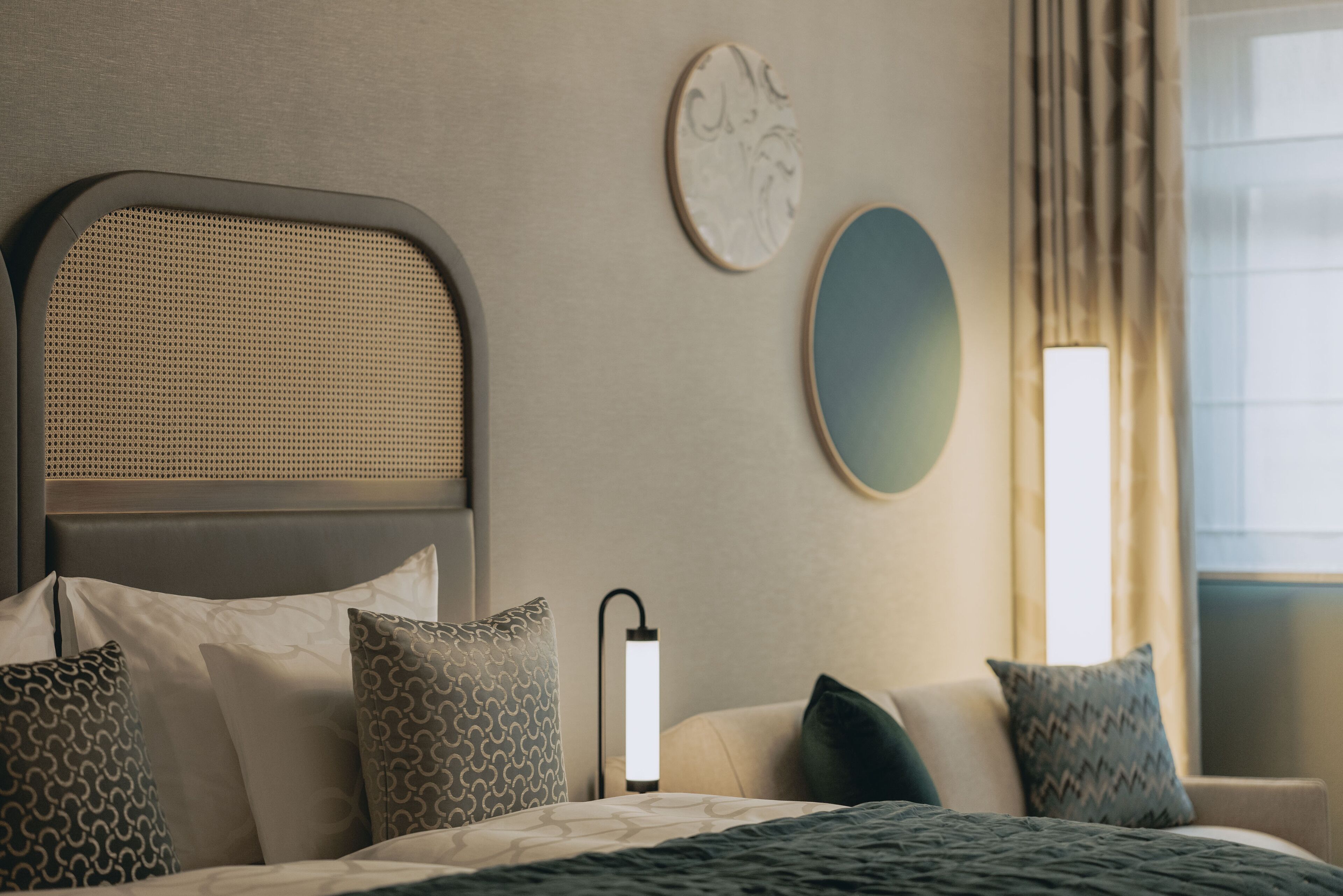 A neatly arranged hotel room with a bed, pillows, a bedside lamp, a wall-mounted mirror, and a window with curtains.