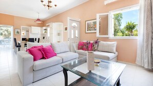 A modern living room with a glass coffee table, two sofas with bright pink and white cushions, a white cabinet, and a view of greenery through a window. - Villa Amorossa with Garden View & Shared Pool by Konnect, Roda Corfu (Corfu)