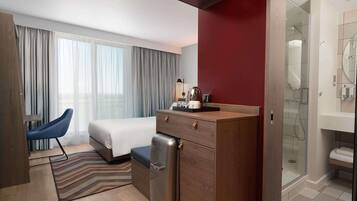 A modern hotel room with a bed, a desk, a refrigerator, a chair, a shower, and a large window with curtains.