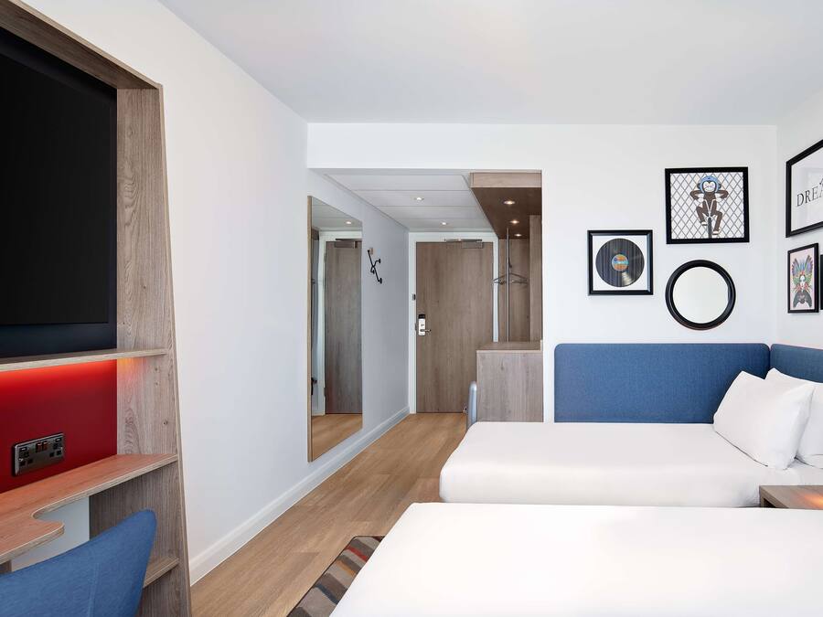 A modern hotel room with a large flat-screen TV, a blue sofa, a white bed, and a wooden accent wall.