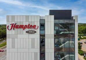 A modern Hampton by Hilton hotel building with a prominent sign, large windows, and a visible staircase.
