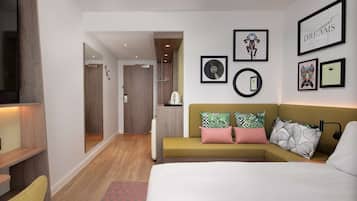 A modern hotel room with a bed, a sofa, a wooden floor, and a hallway leading to a bathroom.