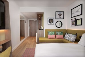 A modern hotel room with a bed, a sofa, a wooden floor, and a hallway leading to a bathroom.