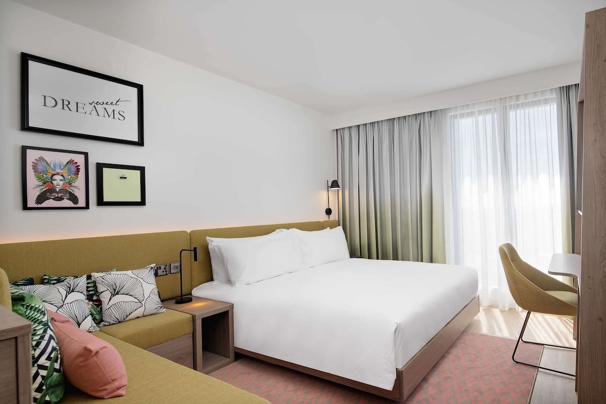 A hotel room with a bed, a bedside table, a lamp, a chair, and a wall with framed pictures.