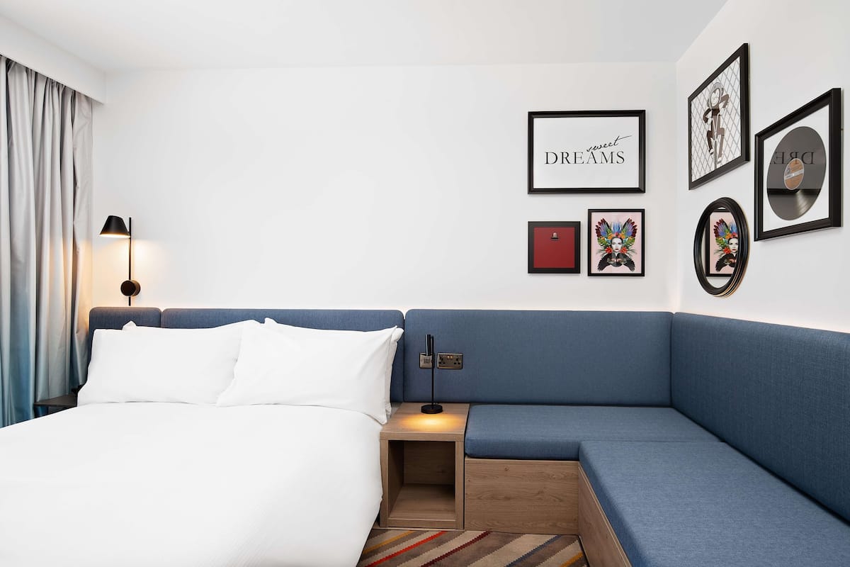 A modern hotel room with a bed, a blue sofa, a nightstand with a lamp, and wall art including a framed picture with the word "DREAMS".