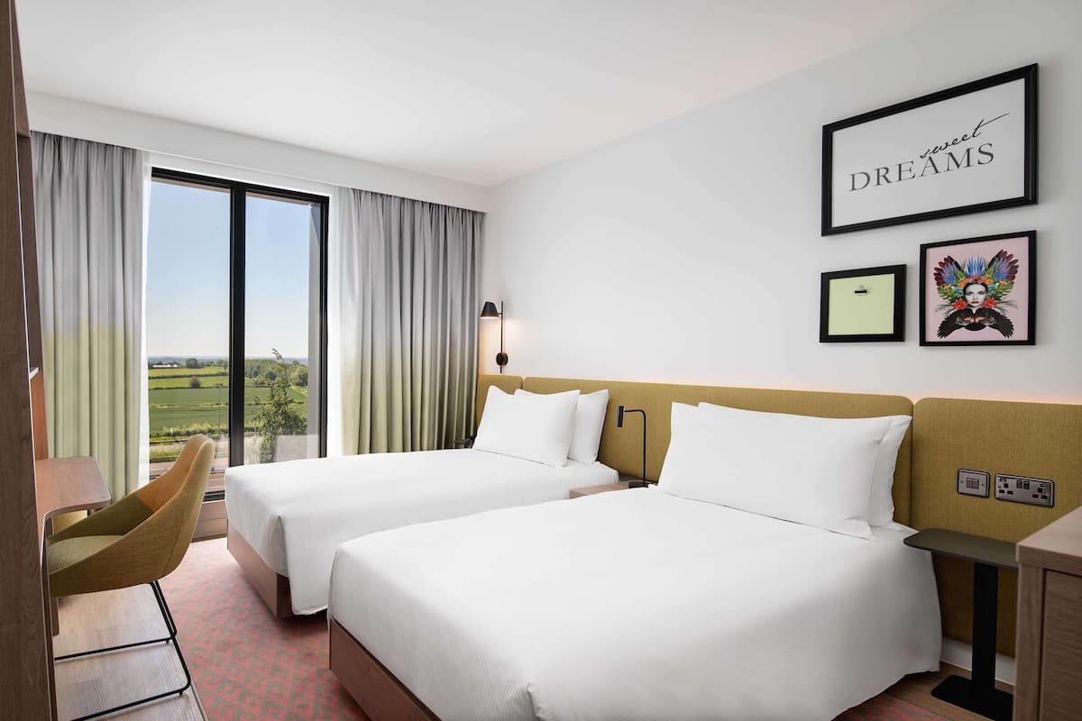 A hotel room with two beds, a desk, a chair, and a large window with curtains.