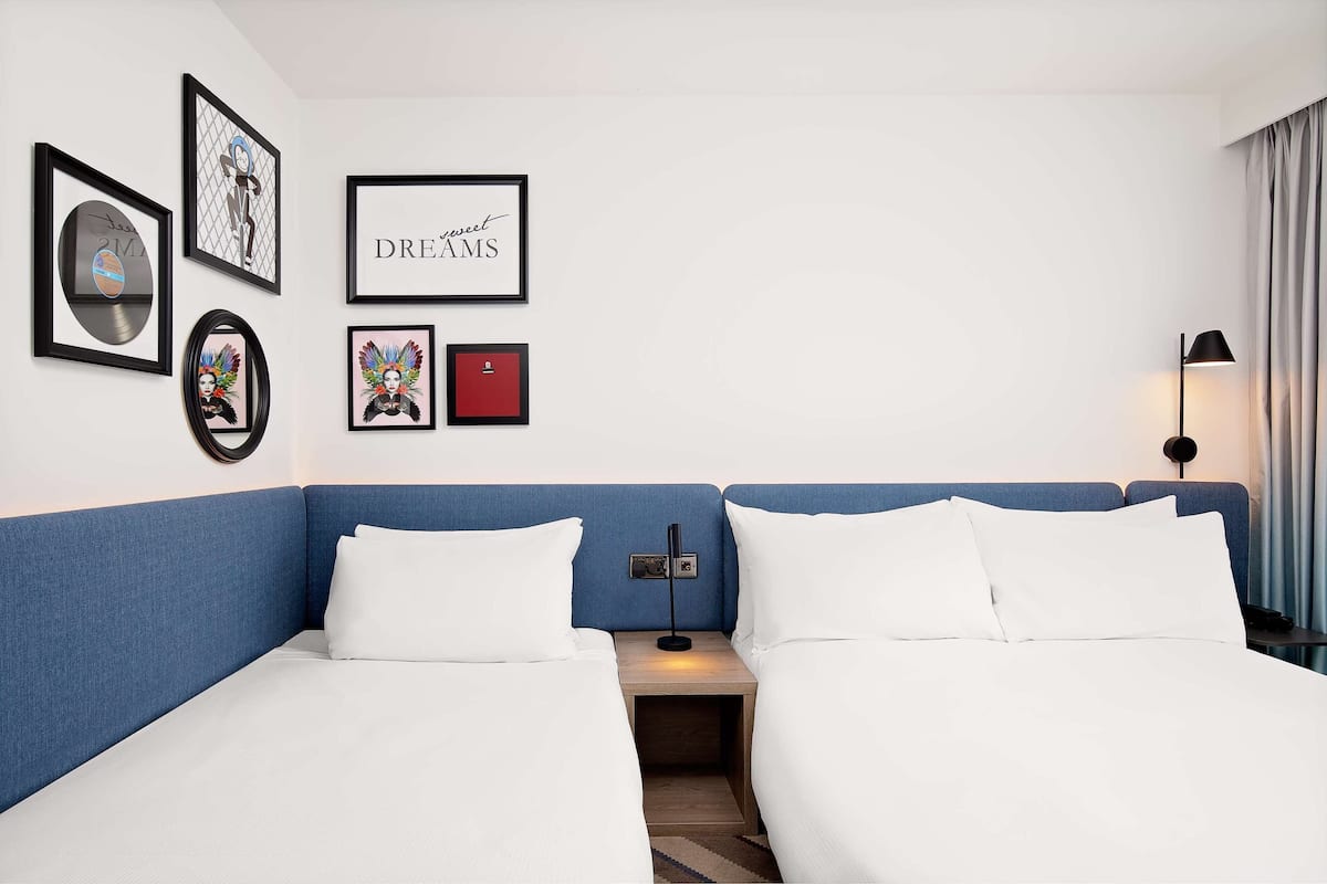 A hotel room with a blue headboard, two beds with white linens, a bedside table with a lamp, and framed artwork on the wall.