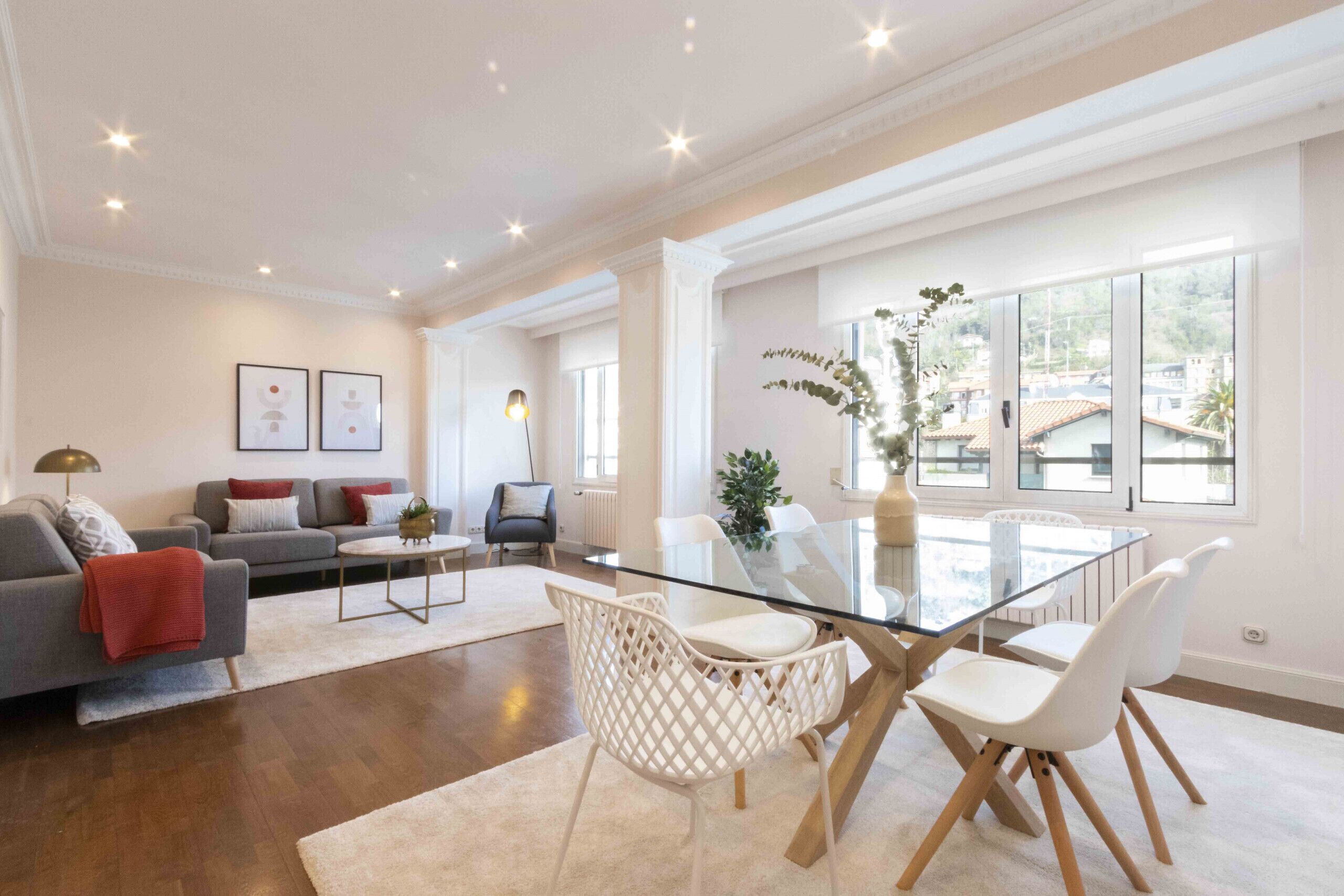 A spacious living room with a glass dining table, white chairs, a grey sofa, and a red throw blanket.