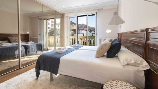 A bedroom with a large mirror, a bed with pillows, a bedside table, and a view of the outside through a sliding door. - Elkano | The Rentals Collection (San Sebastián/Donostia)