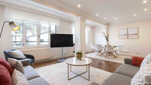 A spacious living room with a large flat-screen TV, comfortable seating, a round coffee table, and a dining area with white chairs. - Elkano | The Rentals Collection (San Sebastián/Donostia)