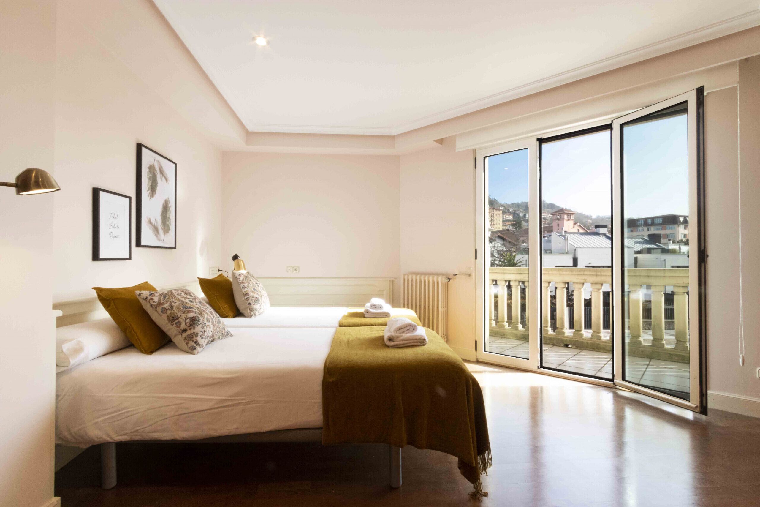 A bedroom with two beds, a large window with a view of buildings, and a radiator on the wall.