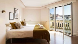 A bedroom with two beds, a large window with a view of buildings, and a radiator on the wall. - Elkano | The Rentals Collection (San Sebastián/Donostia)