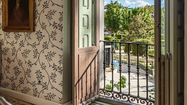A room with a balcony, a framed portrait on the wall, and a patterned wallpaper.
