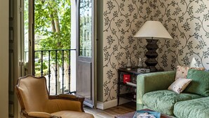 A room with a wooden floor, a green sofa, a brown armchair, a table with a book, and a lamp. There is a window with a view of trees and a balcony.