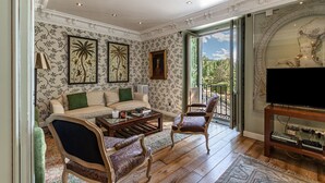 A room with a sofa, armchairs, a coffee table, and a TV. There is a balcony with a view of greenery.