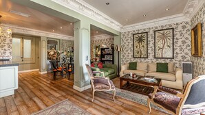 A spacious living room with a large sofa, two armchairs, a coffee table, and a fireplace. The room has wooden flooring, floral wallpaper, and a decorative ceiling.