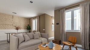 Una sala moderna con un sofá color beige, una mesa de centro de madera y una silla de madera. Hay un ventanal con cortinas, una planta sobre una consola blanca y una pared texturizada.
