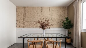 Un comedor con mesa de vidrio, cuatro sillas de madera, un jarrón con flores secas, un muro texturizado y una planta en maceta.