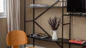 Una sala moderna con una silla de madera, una mesa redonda con revistas y un objeto decorativo, una estantería metálica negra con libros y un cuadro enmarcado, y un televisor sobre un mueble bajo.