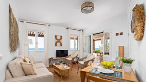 A spacious living room with a sofa, chairs, a TV, and a dining area with a table set for two. - Mon Palau - House with sea view in Biniancolla (Biniancolla)