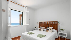A bedroom with a large bed, a wooden headboard, two bedside tables with lamps, a window with curtains, and a view of the sea. - Mon Palau - House with sea view in Biniancolla (Biniancolla)