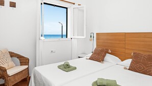 A bedroom with a wooden headboard, a bed with white bedding, a wicker chair, and a window with a view of the sea. - Mercè - House with sea view in Biniancolla (Biniancolla)