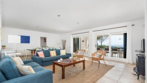 Un vaste espace de vie comprenant un canapé bleu, une table basse en bois et des fauteuils en osier. Une porte vitrée offre une vue sur l’extérieur.