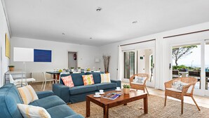 Un salon spacieux avec un canapé bleu, une table basse en bois, des fauteuils en osier et une vue sur l’extérieur grâce à de grandes fenêtres.