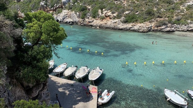Una zona costera con barcos amarrados en un muelle, rodeada de terreno rocoso y vegetación.