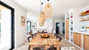 Un comedor amplio con una mesa de madera, sillas y un frutero. Hay lámparas colgantes sobre la mesa y una zona de cocina con armarios y estantes de madera.