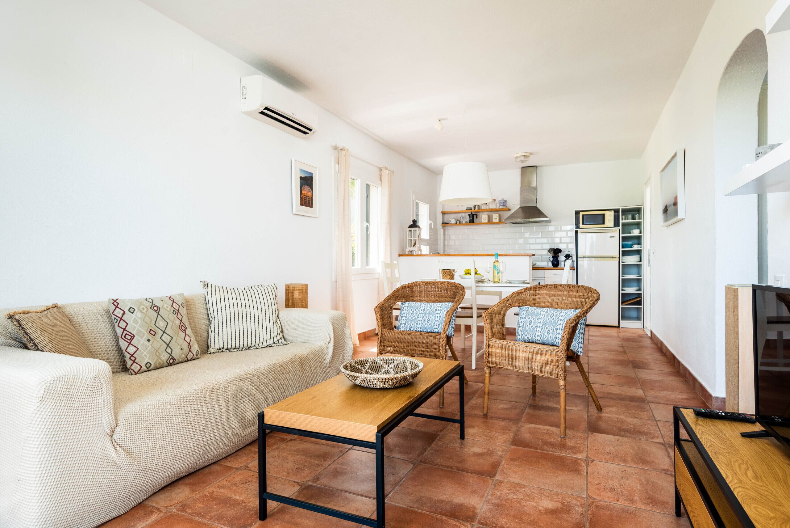 Un salon avec un canapé, une table basse et deux fauteuils en osier. On y trouve également un téléviseur et une télécommande posés sur un meuble en bois.