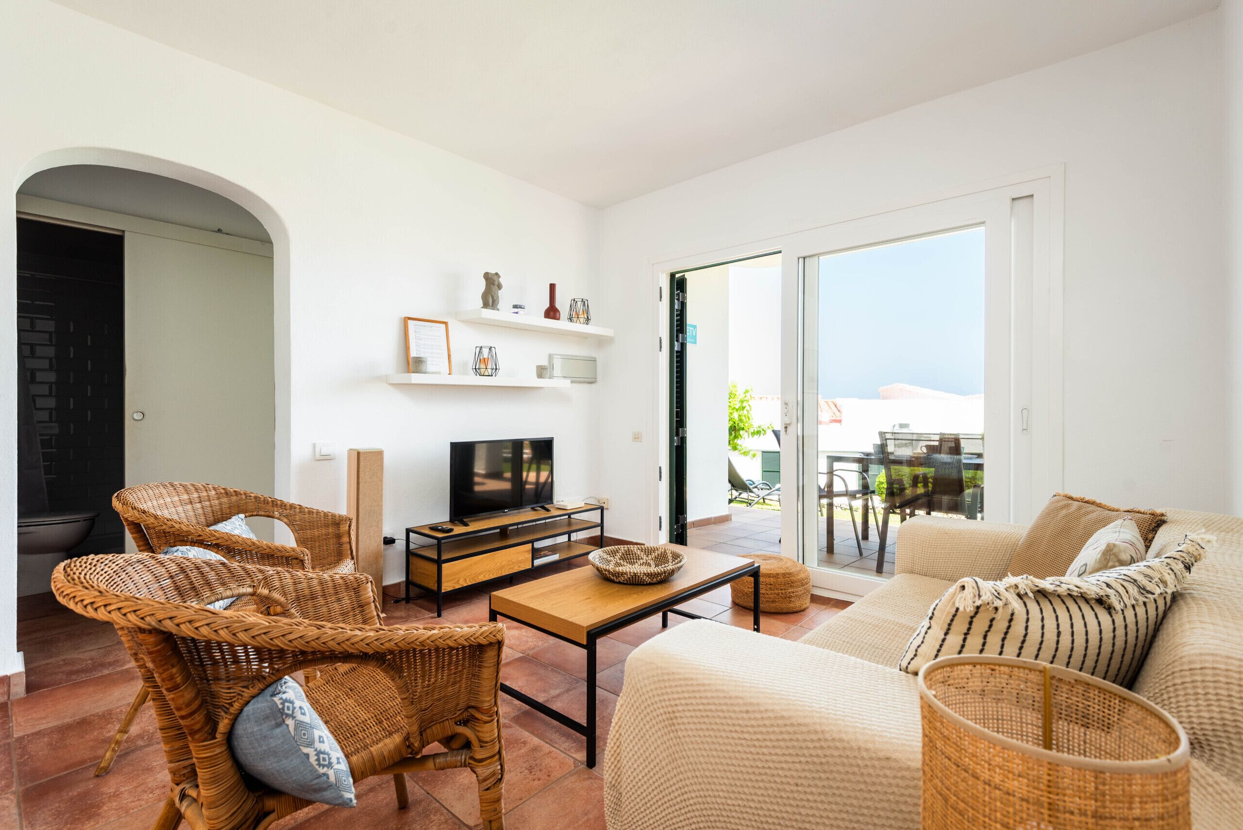 A modern living room with wicker furniture, a flat-screen TV, and a balcony with a view.
