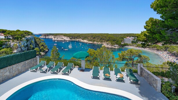 En swimmingpool med solstolar, utsikt över havet med båtar och ett bergigt landskap.
