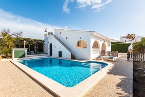 A swimming pool with a white building adjacent, featuring a covered outdoor seating area and a staircase leading to an upper level.