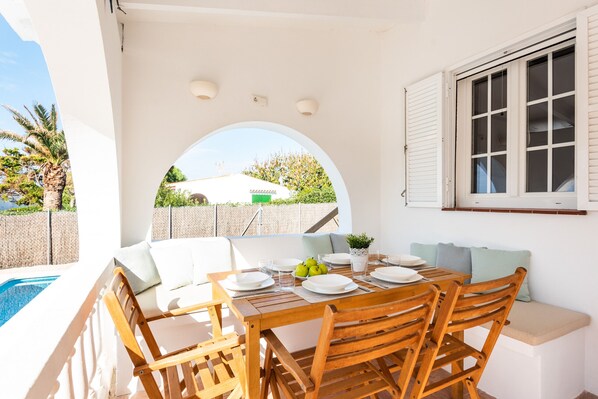 A wooden dining table set with plates, glasses, and a bowl of fruit, surrounded by wooden chairs and cushions, with a view of a swimming pool and greenery outside.